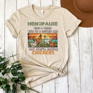 Puede incluir: Camiseta beige con el texto "HENOPAUSE when a woman gets to a certain age and starts buying CHICKENS." El gráfico presenta una escena de puesta de sol con gallinas en un campo. La camiseta tiene las mangas enrolladas.