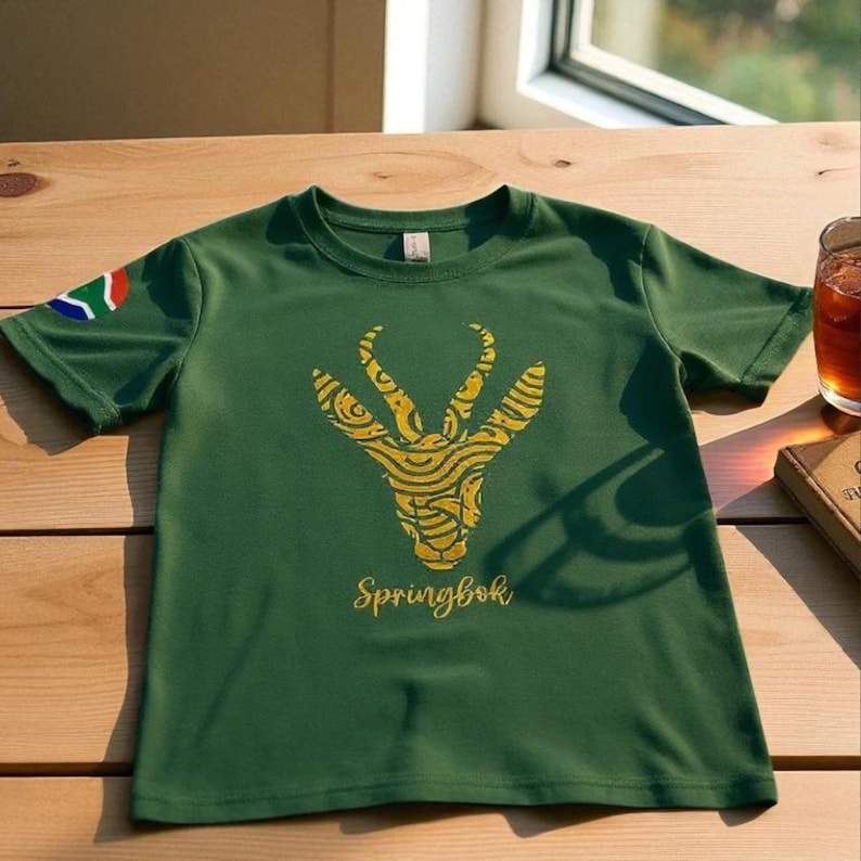 Puede incluir: Camiseta verde con un dise&ntilde;o Springbok dorado y la palabra "Springbok". La bandera de Sud&aacute;frica est&aacute; en la manga izquierda. El dise&ntilde;o es un ant&iacute;lope estilizado. La camiseta est&aacute; sobre una mesa de madera.