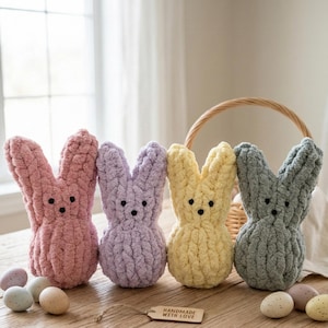 May include: Four handmade, plush bunny figures in pink, lavender, yellow, and gray, with black button eyes. The bunnies are arranged on a wooden surface with decorative Easter eggs and a woven basket in the background. A tag reads "Handmade with Love."