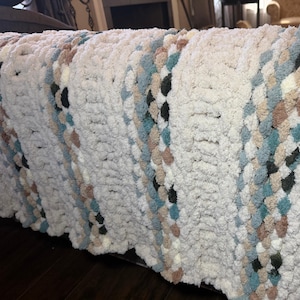May include: A chunky knit blanket in cream with diagonal stripes of teal, brown, and black. The soft, textured blanket is draped over a surface, showcasing its cozy, handmade appearance. This blanket is perfect for adding warmth and style to any room.
