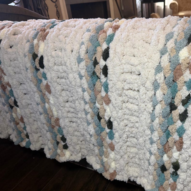 May include: A chunky knit blanket in cream with diagonal stripes of teal, brown, and black. The soft, textured blanket is draped over a surface, showcasing its cozy, handmade appearance. This blanket is perfect for adding warmth and style to any room.