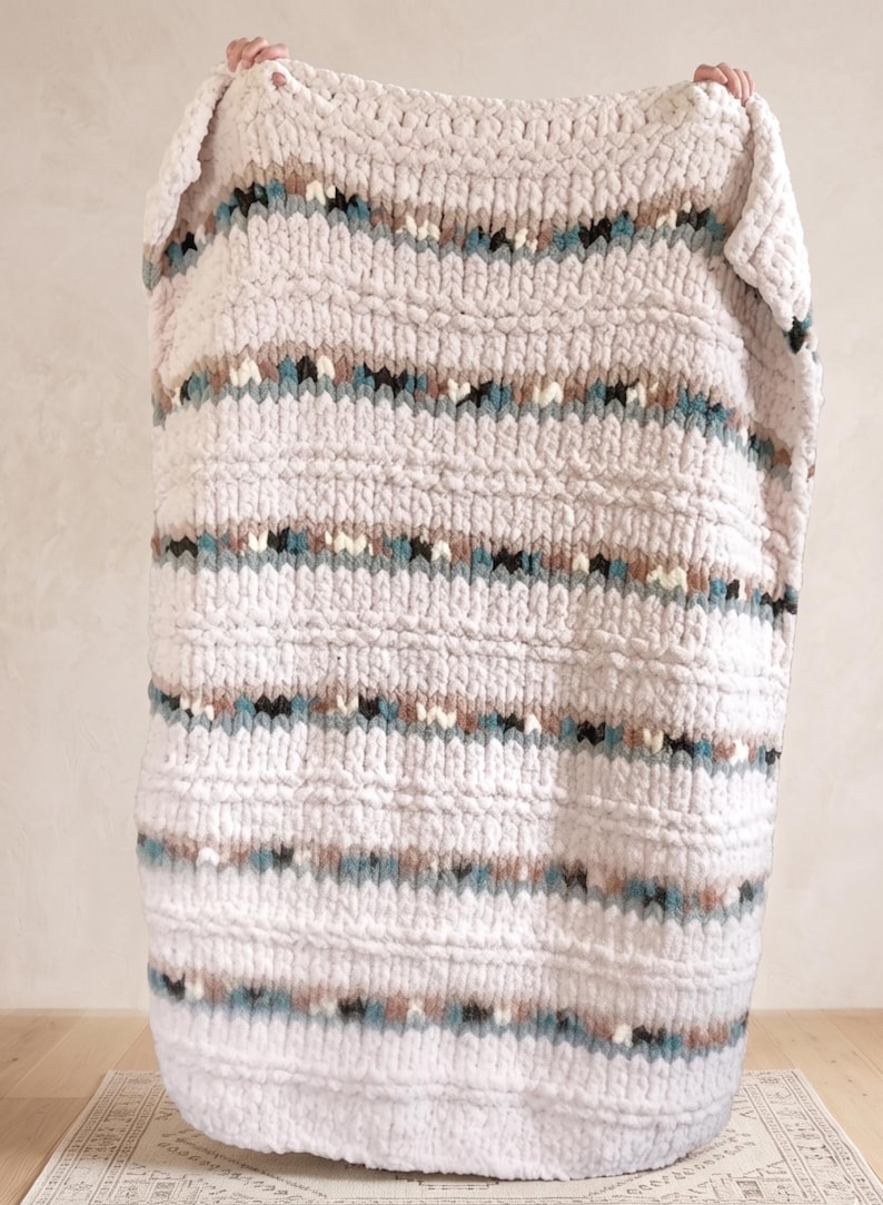 May include: A chunky knit blanket in a light cream color with horizontal stripes of teal, brown, and black. The blanket is made of a soft, textured material, perfect for adding warmth and style to any space.