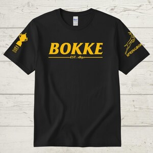 May include: Black t-shirt with gold text and graphics. The word "BOKKE" is centered, with "EST. 1891" below. A trophy graphic and the years 1995, 2007, 2019, and 2023 are on the left sleeve. The right sleeve has a Springbok logo.