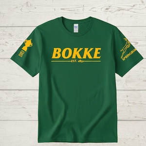 May include: Green t-shirt with the word "BOKKE" in yellow, above "EST. 1891". The left sleeve has a gold trophy graphic with the years 1995, 2007, and 2019. The right sleeve has a yellow Springbok logo.