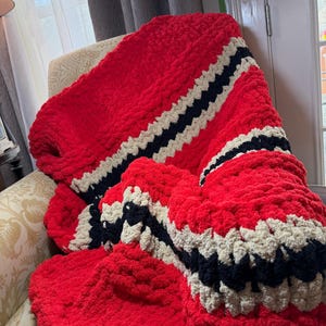 May include: A chunky knit blanket in vibrant red, draped over a chair. The blanket features bold stripes of black and cream, adding a striking contrast to the solid red base. The thick yarn creates a cozy, textured appearance.