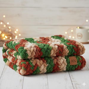 May include: Two folded, chunky knit blankets in shades of green, red, and beige. The blankets are stacked on top of each other, with a small brown leather tag that reads "RN" on the front. The blankets are on a white wooden surface.