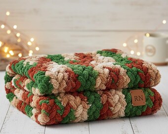 May include: Two folded, chunky knit blankets in shades of green, red, and beige. The blankets are stacked on top of each other, with a small brown leather tag that reads "RN" on the front. The blankets are on a white wooden surface.