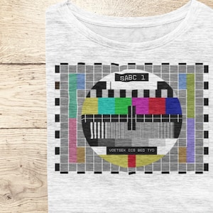 May include: A white t-shirt with a black and white television test pattern graphic. The pattern includes a circle with colored bars and the text "SABC 1" and "VOETSEK DIG BED TVO".