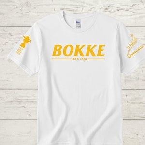 May include: White t-shirt with the word "BOKKE" in yellow block letters, with "EST. 1891" below. A yellow trophy graphic is on the left sleeve, and a Springbok logo is on the right sleeve. The shirt is made of a soft material.