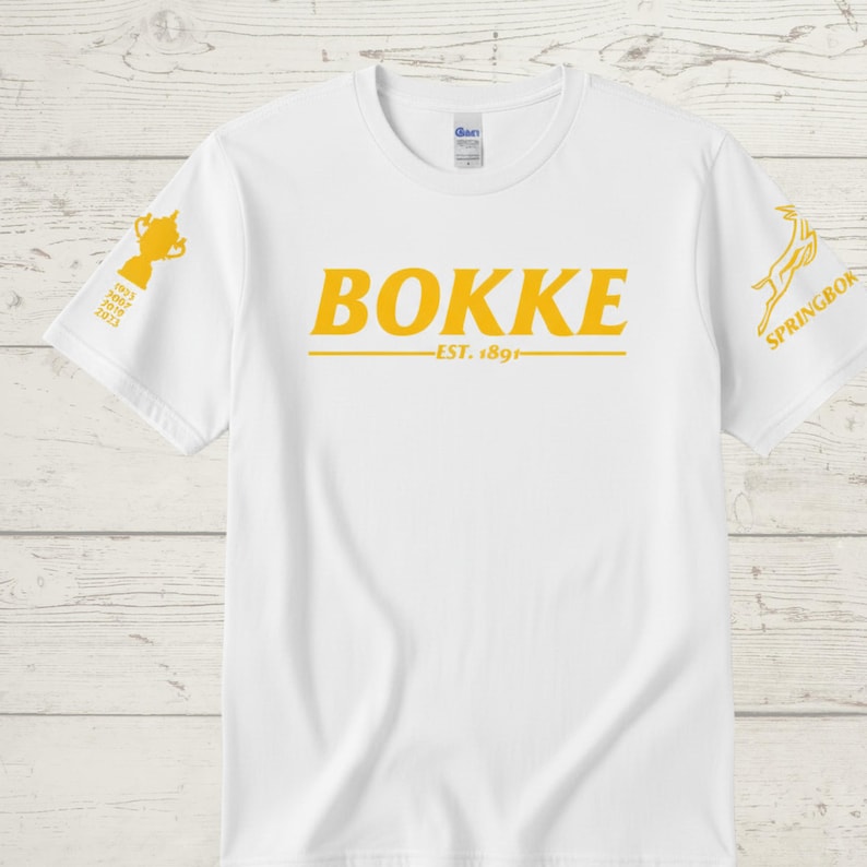 May include: White t-shirt with the word "BOKKE" in yellow block letters, with "EST. 1891" below. A yellow trophy graphic is on the left sleeve, and a Springbok logo is on the right sleeve. The shirt is made of a soft material.
