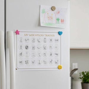 May include: A white refrigerator door displays a weight loss tracker chart with various icons, held by a pink star, blue heart, and yellow smiley face magnets. A child's drawing is pinned above the chart with a gold magnet.