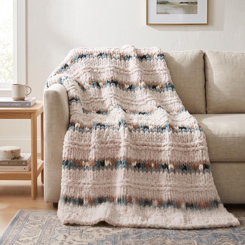 May include: A cream-colored, chunky knit throw blanket with a textured pattern. The blanket features horizontal stripes of blue, brown, and white accents. The blanket is draped over a beige sofa, creating a cozy and inviting aesthetic.