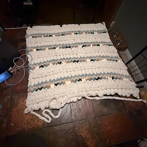 May include: A handmade, chunky knit blanket in cream, with horizontal stripes of teal, brown, and black. The blanket is rectangular and appears soft and cozy, perfect for a bed or sofa. The blanket is lying on a brown tiled surface.