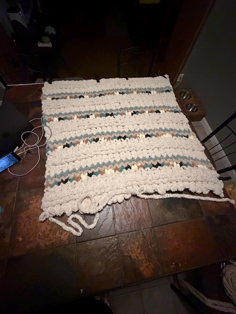 May include: A handmade, chunky knit blanket in cream, with horizontal stripes of teal, brown, and black. The blanket is rectangular and appears soft and cozy, perfect for a bed or sofa. The blanket is lying on a brown tiled surface.