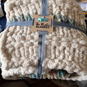May include: A thick, cream-colored blanket with a chunky knit texture, tied with a gray ribbon. The blanket has a decorative border with muted green, pink, and blue hues. A wooden tag reads "The Rustic Blanket."