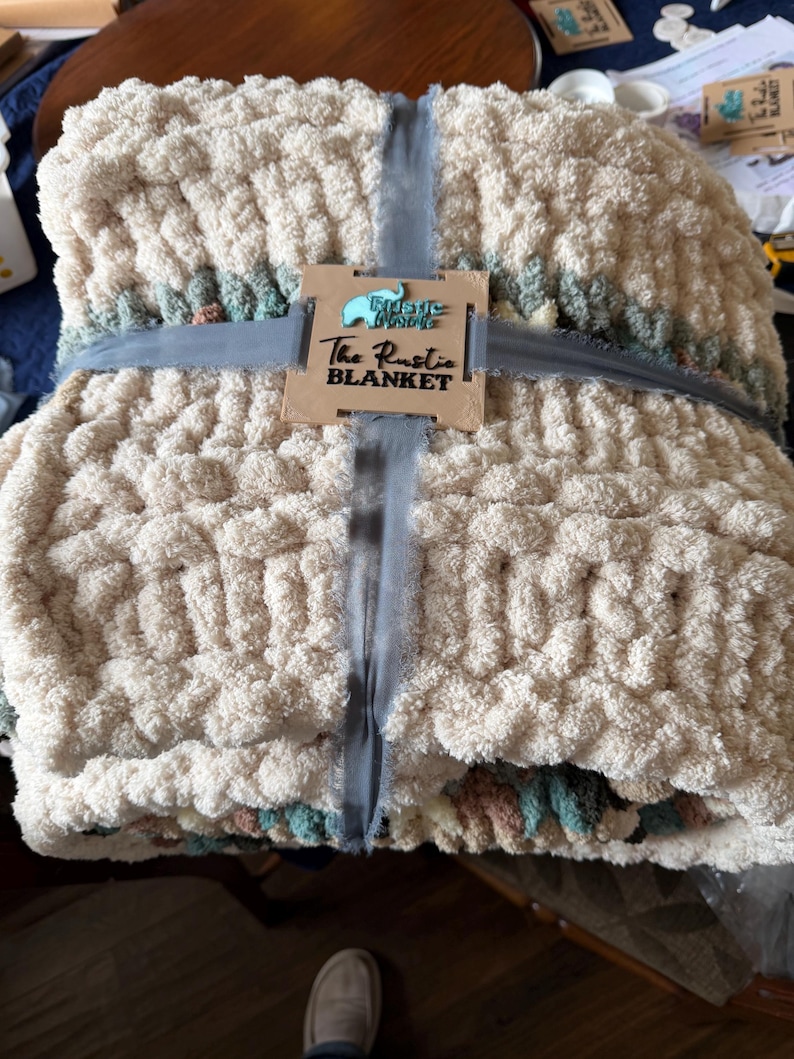 May include: A thick, cream-colored blanket with a chunky knit texture, tied with a gray ribbon. The blanket has a decorative border with muted green, pink, and blue hues. A wooden tag reads "The Rustic Blanket."