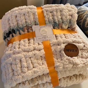 May include: A cream-colored chunky knit blanket, tied with an orange ribbon. The blanket has a textured appearance and a leather patch with the name "Hailey". A tag reads "The Classic Yarn".