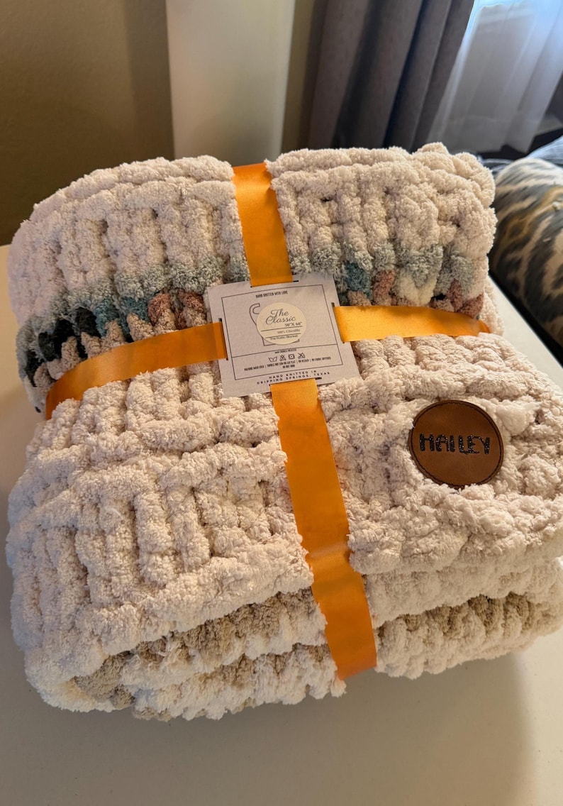 May include: A cream-colored chunky knit blanket, tied with an orange ribbon. The blanket has a textured appearance and a leather patch with the name "Hailey". A tag reads "The Classic Yarn".