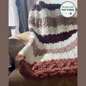 May include: A chunky knit throw blanket featuring horizontal stripes in shades of cream, tan, and dark brown. The textured yarn creates a soft, looped appearance. A circular logo in the upper right corner displays the text Breadcrumb PATTERN.
