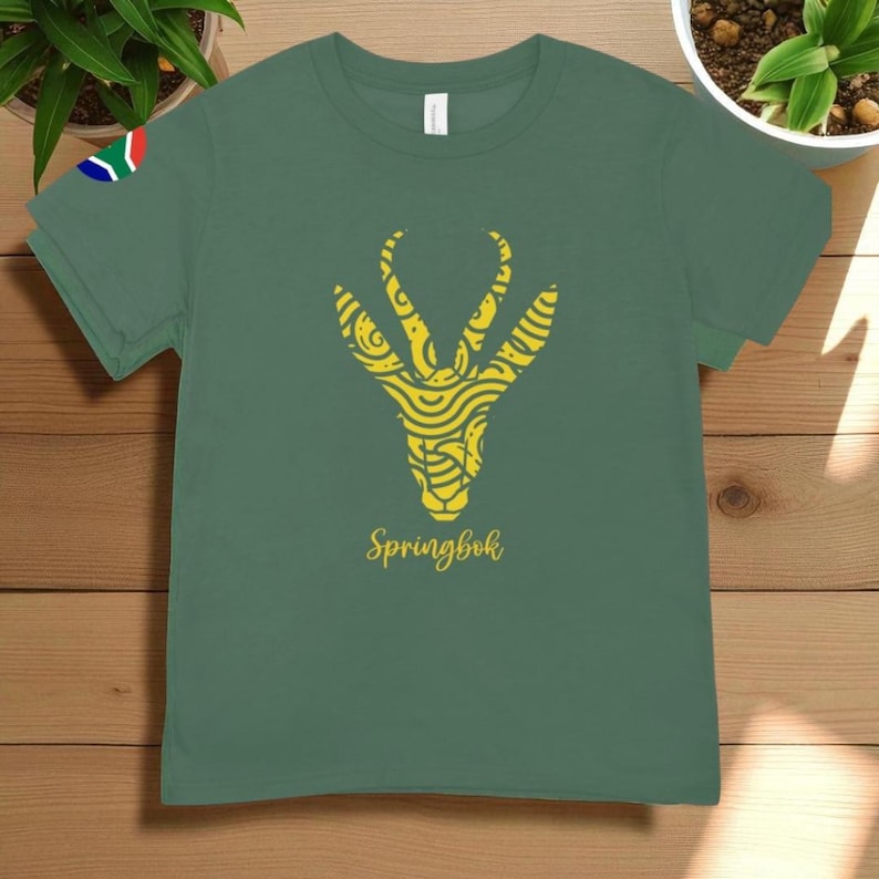 Puede incluir: Una camiseta verde con un dise&ntilde;o de Springbok amarillo. El dise&ntilde;o es una ilustraci&oacute;n estilizada de una cabeza de springbok con patrones detallados. La palabra "Springbok" est&aacute; escrita en cursiva debajo. La bandera sudafricana est&aacute; en la manga izquierda.