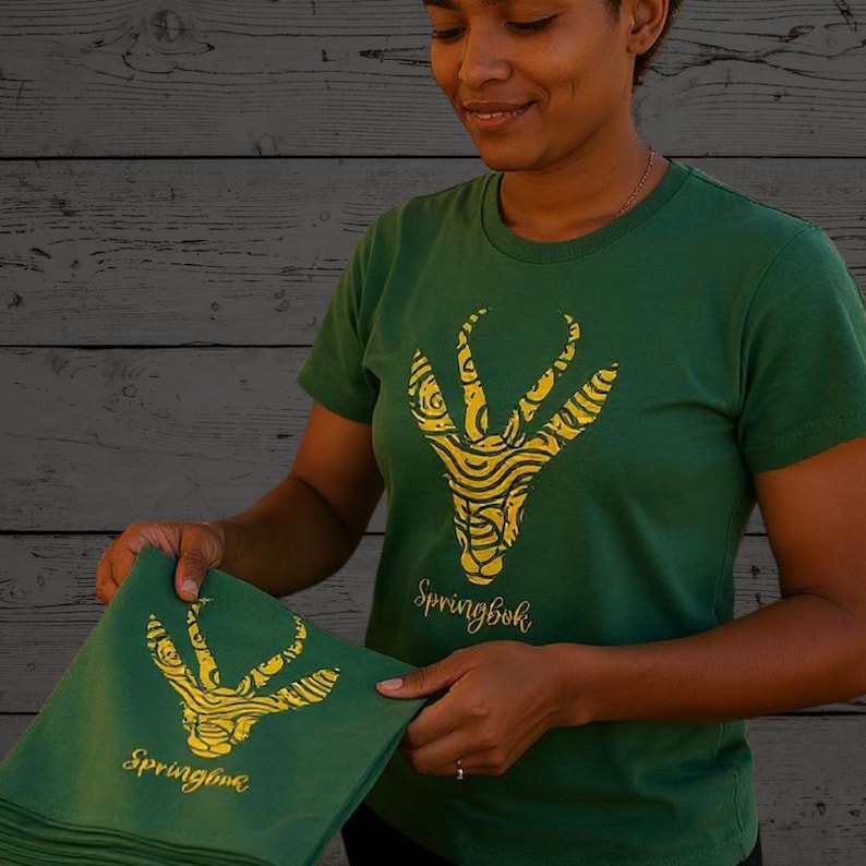 Puede incluir: Camisetas verdes con un dise&ntilde;o dorado de Springbok. El dise&ntilde;o es una ilustraci&oacute;n estilizada de la cabeza y los cuernos de un Springbok. La palabra "Springbok" est&aacute; impresa debajo del dise&ntilde;o en una fuente de escritura. Las camisetas est&aacute;n siendo sostenidas por una persona.
