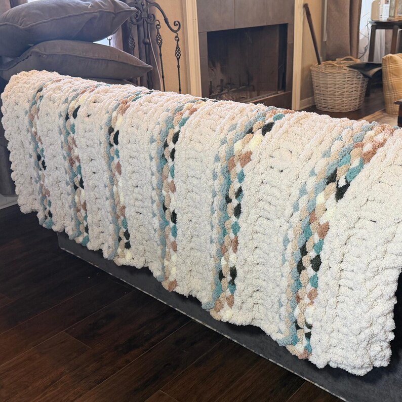 May include: A chunky knit blanket in cream with accent stripes of light blue, tan, and black. The blanket is draped over a dark gray bench, showcasing its textured, cozy appearance. This handmade blanket is perfect for home decor.