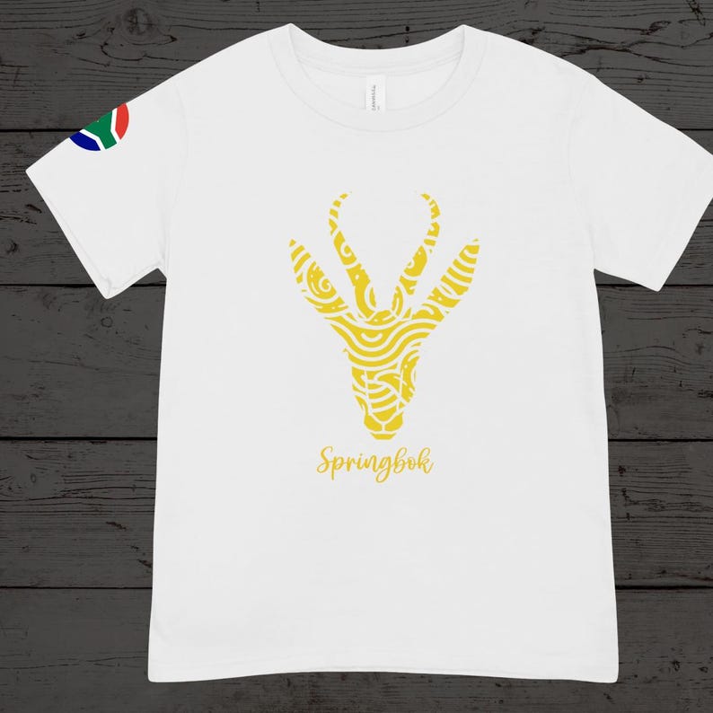 Puede incluir: Camiseta blanca con un dise&ntilde;o de cabeza de Springbok amarilla con patrones intrincados. La palabra "Springbok" est&aacute; escrita debajo. La bandera sudafricana est&aacute; en la manga izquierda.