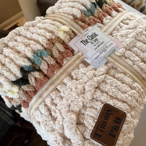May include: An XL-sized, cream-colored chunky knit blanket with a woven design. The blanket is tied with a beige ribbon and features a tag that reads "The Classic". A brown leather patch with the text "Timmy PhD" is attached to the blanket.