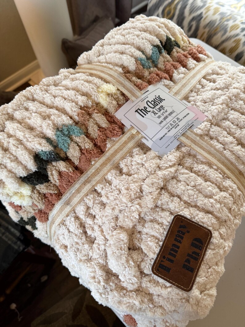 May include: An XL-sized, cream-colored chunky knit blanket with a woven design. The blanket is tied with a beige ribbon and features a tag that reads "The Classic". A brown leather patch with the text "Timmy PhD" is attached to the blanket.