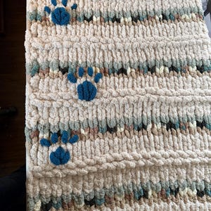 May include: A cream-colored, chunky knit blanket with three blue paw prints. The blanket has horizontal stripes of various colors, including blue, brown, and green. The texture appears soft and plush, suitable for pets.