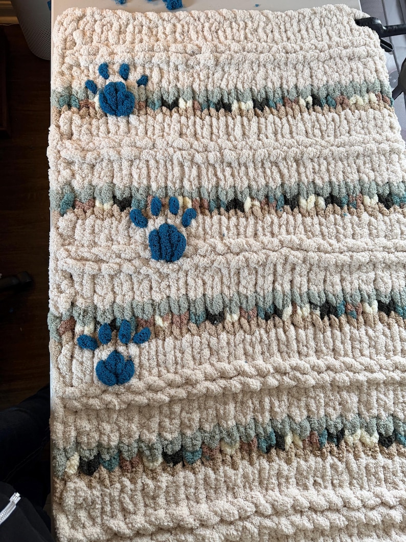 May include: A cream-colored, chunky knit blanket with three blue paw prints. The blanket has horizontal stripes of various colors, including blue, brown, and green. The texture appears soft and plush, suitable for pets.