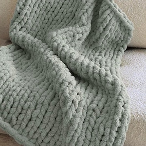 May include: A sage green chunky knit blanket featuring a thick, hand-knitted texture with large, soft yarn loops. The blanket is draped in folds, showcasing the consistent, interlocking stitch pattern throughout the entire piece.