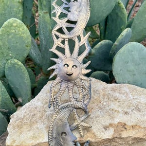 Puede incluir: Un colgante de pared de metal con diseños celestiales. La obra de arte incluye un sol sonriente, lunas crecientes y estrellas. La pieza está hecha de metal de color plateado y se exhibe sobre un fondo de piedra natural y cactus.