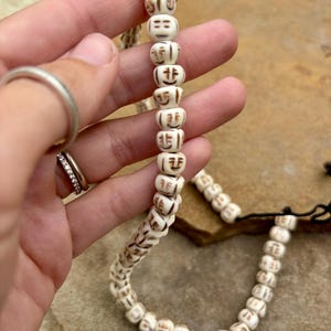 Op de afbeelding: Crèmekleurige kralenketting met herhaalde gesneden gezichtsontwerpen. De kralen zijn geregen met een zwart koord. De ketting wordt in een hand gehouden en toont de ingewikkelde details van de gesneden gezichten.
