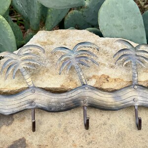 May include: Metal wall hook with a tropical theme. The hook features a wavy design with five palm trees and four hooks. The metal has a distressed, weathered appearance, with shades of silver and bronze. The hook is mounted on a stone.