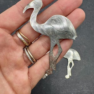 May include: A silver-toned flamingo brooch with detailed feather and leg designs. The brooch is held in a hand, showcasing its intricate craftsmanship. A smaller, matching flamingo piece is also visible.