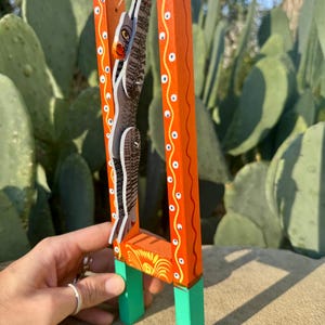 May include: A hand-painted wooden sculpture featuring a stylized figure within an orange frame. The frame has white and yellow decorative accents. The figure is primarily black and white with a red and yellow face, and green feet.