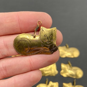 May include: A set of seven gold-toned metal cat charms. Each charm features a detailed etched design of a cat lying down. The charms have a loop at the top for attaching to jewelry or other crafts.