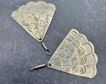 May include: Two silver-colored fan-shaped earrings with intricate engraved designs. Each earring has a coiled wire and a small loop for attachment. The earrings are displayed on a dark gray surface.