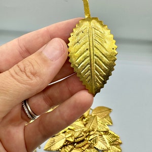 May include: A pile of gold-colored metal leaf charms with a single leaf charm held in a hand. The leaf charm has a hole at the top for hanging.