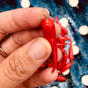 May include: A close-up of a bright red chili pepper charm with a gold loop at the top. The charm is being held by a hand, and there are other red chili pepper charms in the background.