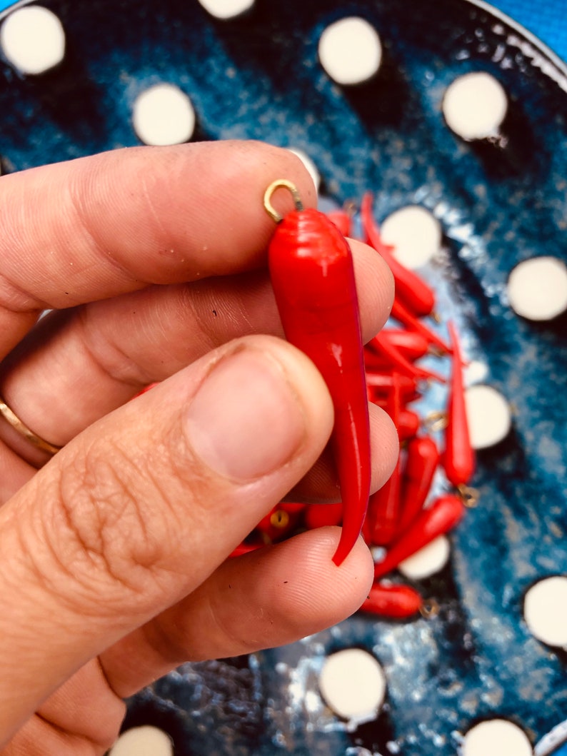 May include: A close-up of a bright red chili pepper charm with a gold loop at the top. The charm is being held by a hand, and there are other red chili pepper charms in the background.
