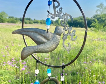 May include: A metal wind chime featuring a dove and the word "Peace" within a circular frame. The chime is adorned with blue glass beads and bells, hanging against a backdrop of wildflowers.