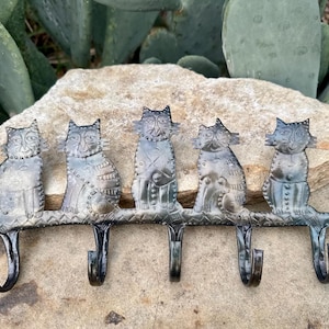Puede incluir: Un colgador de pared de metal con cinco figuras de gatos. Los gatos están posados en una barra horizontal con cinco ganchos debajo. El metal tiene un aspecto envejecido y desgastado. El fondo incluye una roca y plantas verdes.
