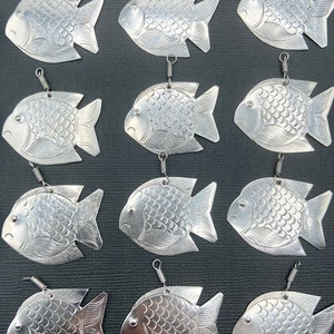May include: Twelve silver fish-shaped ornaments with a loop at the top for hanging. Each ornament has a textured surface resembling fish scales.
