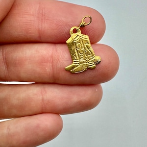 May include: Gold-toned metal charm shaped like a cowboy boot with intricate details. The boot has a spur and a decorative design on the shaft.