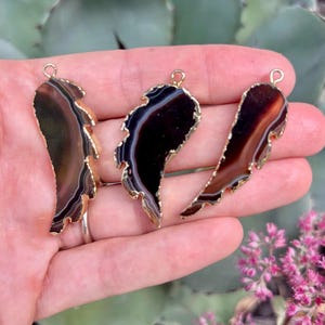 May include: Three irregular, wing-shaped agate pendants featuring dark brown and black banded patterns with translucent layers. Each stone is edged in a metallic gold finish and includes a small circular gold-toned metal loop at the top for hanging.