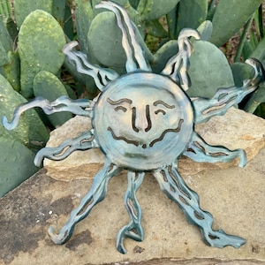 May include: A metal sun sculpture with a smiling face and wavy rays. The sun has a blue-green patina with bronze highlights. The sculpture is placed on a rock with a cactus in the background.