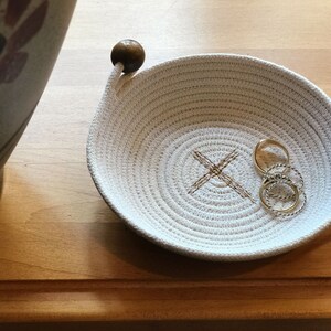 May include: A white cotton rope bowl with a wooden handle and a brown X stitched into the center. Three gold rings are inside the bowl.