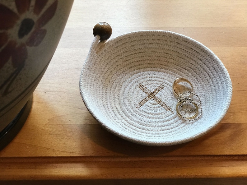 May include: A white cotton rope bowl with a wooden handle and a brown X stitched into the center. Three gold rings are inside the bowl.